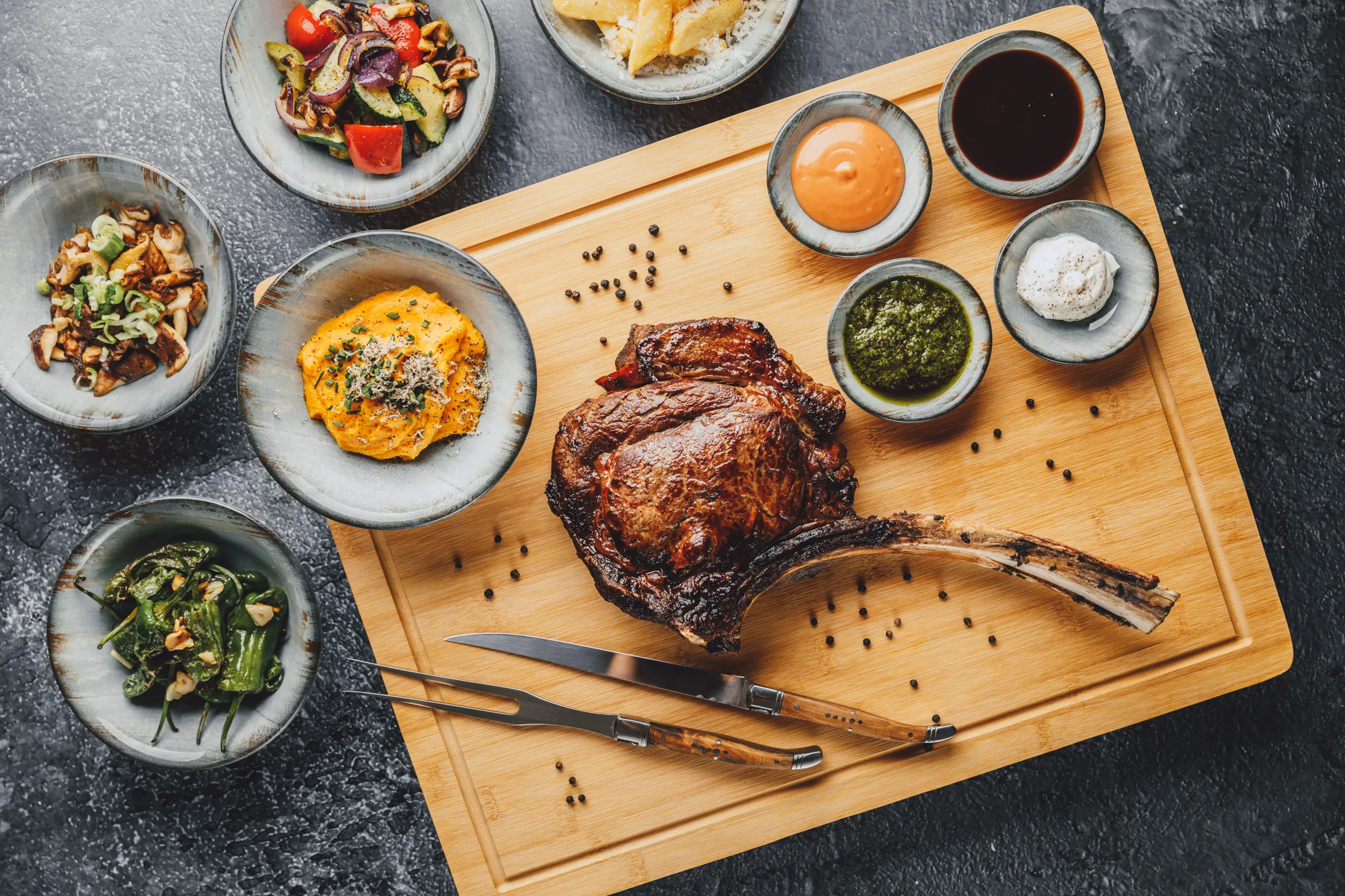 Tomahawk-Steak auf Holzbrett mit Beilagen und Saucen von oben, Das Brixx