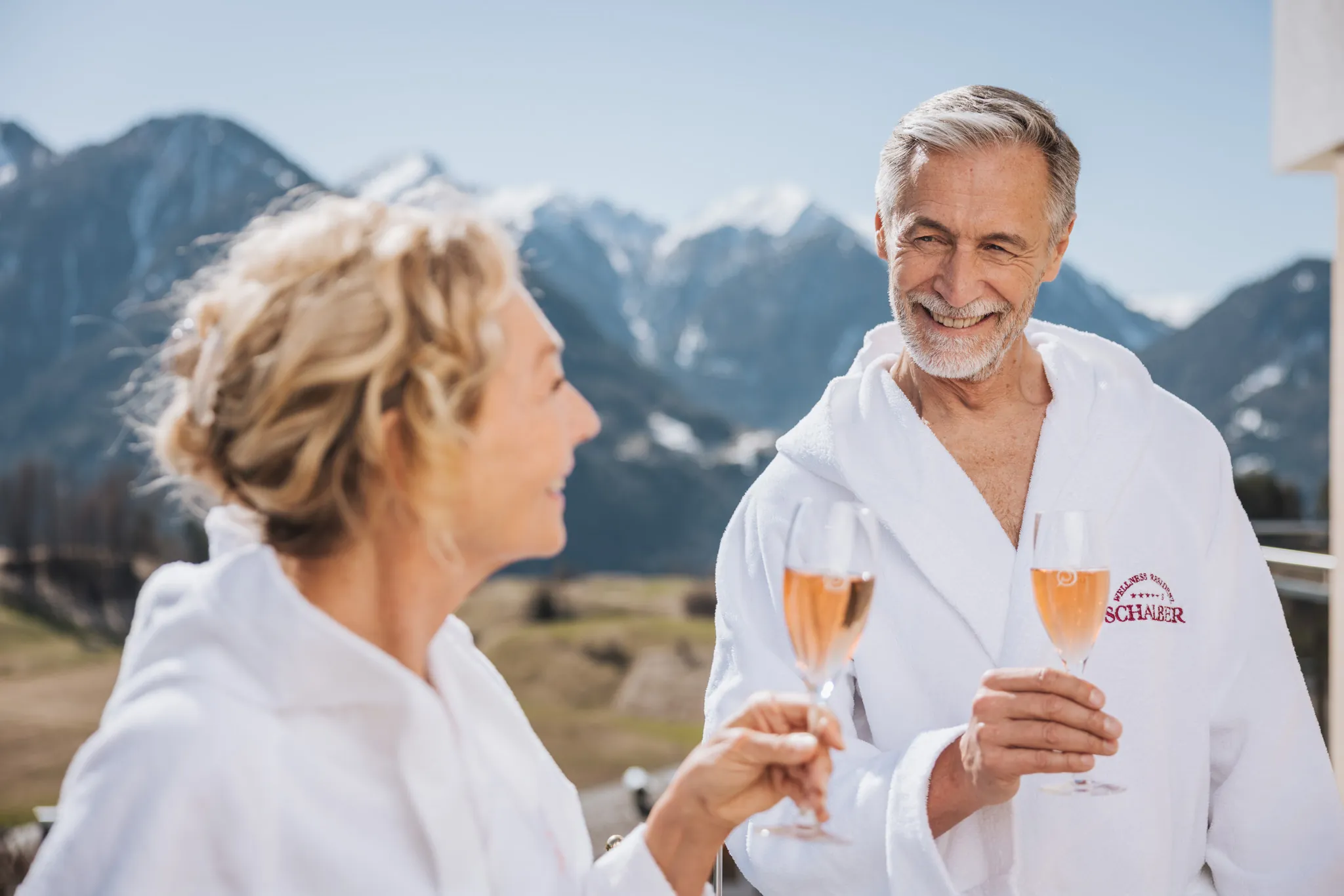 Paar im Bademantel stößt mit Prosecco vor dem Bergpanorama an, Wellnessresidenz Schalber